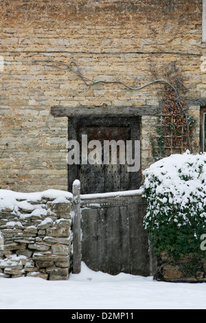 En pierre de Cotswold cottage au toit de chaume avec porte avant en bois, jardin mur et porte en bois. La neige tombe et se pose sur le sol. Banque D'Images