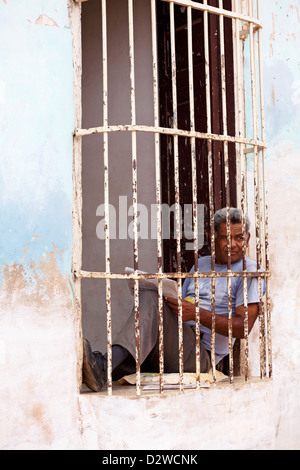 La vie quotidienne à Cuba - homme assis sur coussin dans la fenêtre avec du papier journal derrière les barreaux à Trinidad, Cuba Banque D'Images