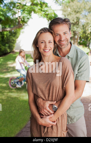 Portrait of smiling couple hugging in allée avec sa fille en bicyclette en arrière-plan Banque D'Images