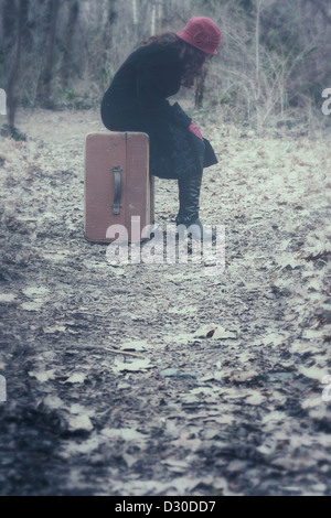 Une femme avec un manteau noir et un chapeau rouge est assis sur une vieille valise dans les bois Banque D'Images