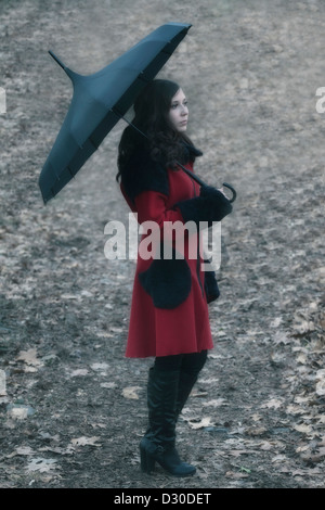 Une femme avec un manteau rouge et un parapluie marche sur une piste forestière Banque D'Images