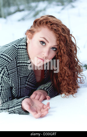 Belle jeune femme aux cheveux rouges et bouclés noir et blanc manteau herringbone réside sur l'estomac dans la neige Banque D'Images