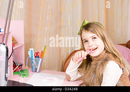 Jolie petite fille heureux assis à son bureau de mâcher son crayon rose à la recherche jusqu'à la rêverie de plafond avec un beau sourire sur son visage. Banque D'Images