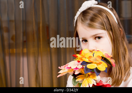 Jolie petite blonde timide fille avec un bouquet de fleurs en les tenant devant le visage comme elle regarde la caméra contre un rideau toile avec copyspace. Banque D'Images