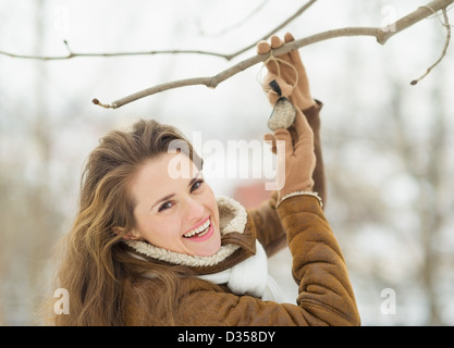 Smiling young woman hanging mangeoire sur arbre dans winter park Banque D'Images