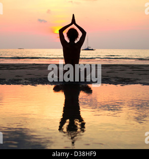 Yoga woman sitting in lotus pose sur la plage pendant le coucher du soleil, avec la réflexion dans l'eau