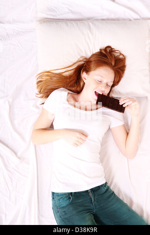 Une jeune femme rousse de manger du chocolat au lit. Banque D'Images