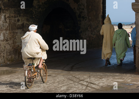 Homme marocain dans un jalabiya vélo dans une des anciennes portes d'entrée du port d'Essaouira, Essaouira, Maroc Banque D'Images