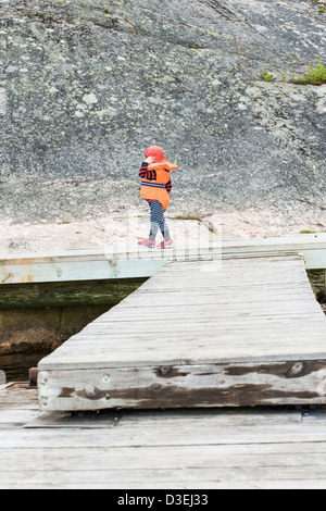 Jeune enfant portant un gilet de marcher sur la jetée en bois avec des pierres à l'arrière-plan Banque D'Images