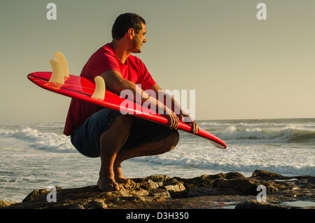 Un surfeur en regardant les vagues s'asseoir avec ses bras autour de son surf. Banque D'Images