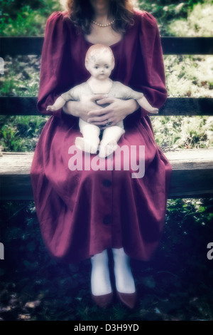 Une femme en robe rouge est assise sur un banc en bois avec une vieille poupée sur ses genoux Banque D'Images