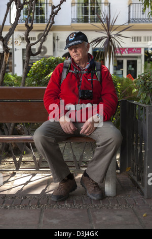 Senior male avec un appareil photo autour du cou et un sac à dos sonner pendant sur un banc de parc Banque D'Images