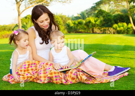 Photo de jeune mère avec deux enfants mignons livre de lecture en plein air au printemps, heureuse maman de ses enfants l'enseignement dans le parc, Banque D'Images