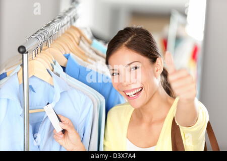 Portrait of happy Young / Chinois asiatique multiethnique young woman showing Thumbs up tout en maintenant l'étiquette de prix sur les vêtements au shop Banque D'Images