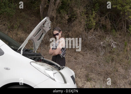 Jeune femme adulte avec bras croisés l'air, avec anxiété, sa voiture en panne avec le capot en place. Banque D'Images