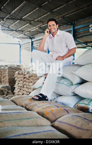 Homme assis sur pile de sacs de blé et de parler sur un téléphone mobile dans un entrepôt, Anaj, Mandi, Sohna Gurgaon, Haryana, Inde Banque D'Images