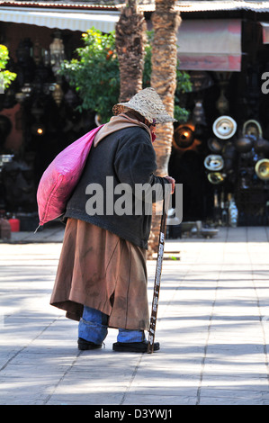 Vieil homme avec chapeau de paille, bâton de marche et rose sac sur son épaule en traversant une place de la ville de Marrakech, Maroc Banque D'Images