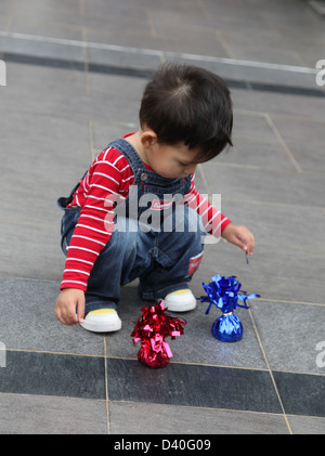 C'est une photo d'une petite Asian kid enfant garçon qui joue avec 2 petit sachet sur le carrelage. Il porte des rayures rouge et blanc Banque D'Images