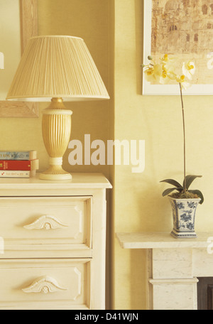 Chambre jaune pâle avec crème lampe avec abat-jour plissé sur la poitrine-de-tiroirs à côté de cheminée avec des orchidées en pot Banque D'Images