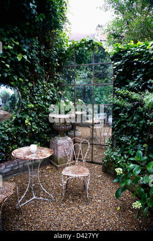 Vieux métal table et chaises sur le gravier en face de miroir en forme de fenêtre en arc au townhouse jardin avec des murs couverts de lierre Banque D'Images