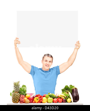 Beau Jeune homme tenant un panneau vierge et posant avec un tas de fruits et légumes frais isolé sur fond blanc Banque D'Images