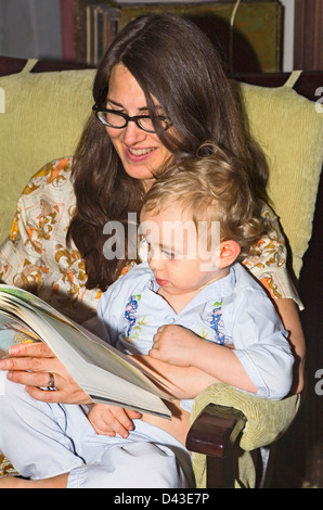 Maman aux cheveux noirs se trouve dans la lecture de livre président blonde pré-scolaire fils en pyjama en soir Banque D'Images