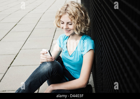 Portrait of Teenage Gril souriant, assis sur le trottoir à l'aide de téléphone cellulaire, Mannheim, Allemagne Banque D'Images