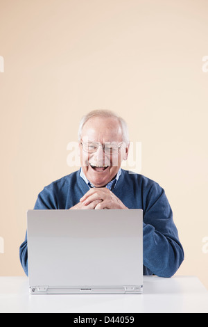 Senior Man sitting at Table looking at laptop computer rire, Studio Shot sur fond beige Banque D'Images