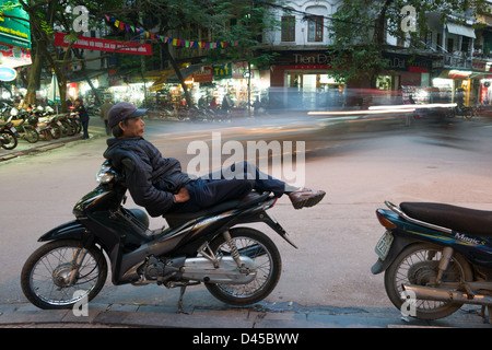 Moto taxi chauffeur de vous détendre sur sa moto que le trafic passe sur Lo Su, dans le vieux quartier de Hanoi, Vietnam Banque D'Images