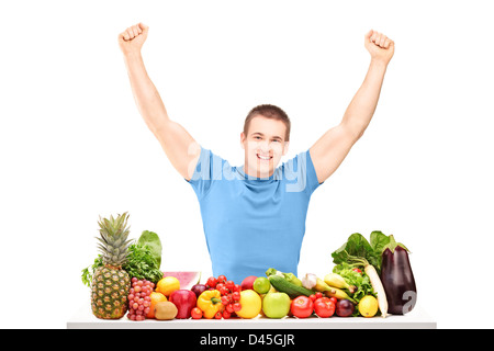 Guy excité gesticulant bonheur assis sur une table pleine de fruits et légumes frais isolé sur fond blanc Banque D'Images