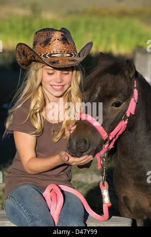 Très jolie jeune femme alimente un traiter à son cheval miniature, Portrait of a smiling girl wearing a cowboy hat. Banque D'Images