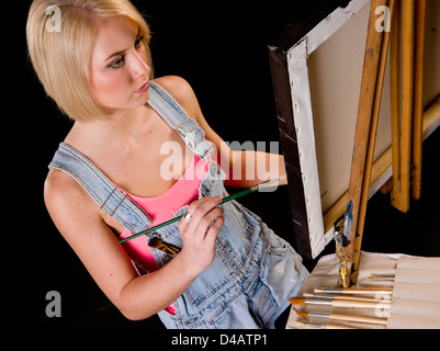 Une bonne femme blonde habillée dans des combinaisons fournit des touches de finition à la peinture de chevalet sur Banque D'Images