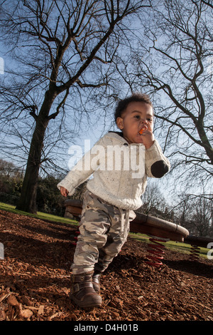 Mixed Race Bébé garçon dans l'alimentation du parc à pied orange avec des arbres derrière le port de cavalier à la crème Banque D'Images