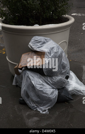 Homme sans foyer enveloppé de mendicité et simultanément la lutte contre la pluie, Lexington Ave., Manhattan. Banque D'Images