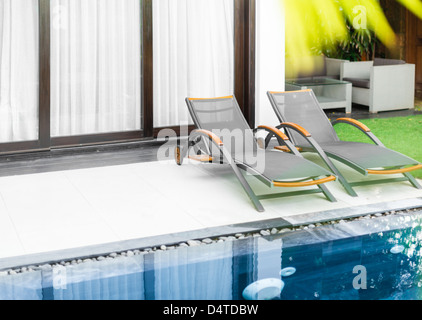 Deux transats sur sol blanc et fenêtres avec rideaux derrière. Pelouse verte, une table et un fauteuil en arrière-plan. Chambre De luxe avec piscine Banque D'Images