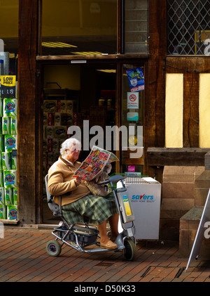 Femme âgée en scooter de mobilité lire un journal à l'extérieur d'un magasin à Stafford Angleterre UK Banque D'Images