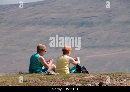 Vue arrière de 2 femmes ou femelles (marcheurs) assis sur le sommet de l'ensoleillée le Pen-y-ghent, manger, pique-nique au-delà fellside raide - Vallées du Yorkshire, England, UK. Banque D'Images