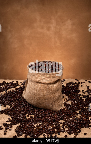 Un sac de jute rempli de grains de café et entouré de haricots renversé contre un fond brun, marbrée. Banque D'Images