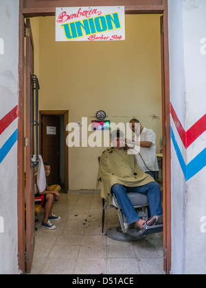 Un salon de coiffure coupe un homme d'âge moyen dans les cheveux de son salon de coiffure chaise tandis qu'un jeune garçon attend d'avoir couper les cheveux à La Havane, Cuba Banque D'Images