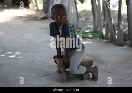 Un jeune garçon africain assis sur un tabouret au milieu d'un village. Banque D'Images