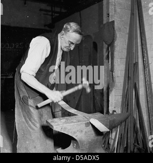 Historique, années 1950. Forgeron âgé portant un long tablier et dans un gilet de costume utilisant une enclume et un marteau pour mouler un long morceau de métal. Banque D'Images