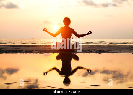 Yoga woman sitting in lotus pose sur la plage pendant le coucher du soleil, avec la réflexion dans l'eau, dans des couleurs vives.