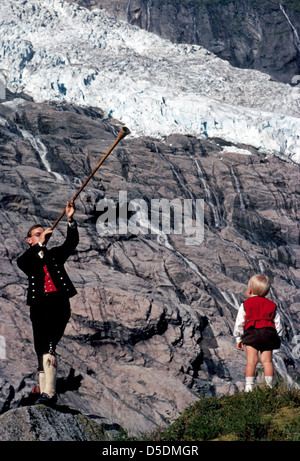 Un jeune homme norvégien en vêtements traditionnels d'antan souffle un cor des alpes en bois vintage pour son fils sous un glacier dans l'Fjaerland, la Norvège. Banque D'Images