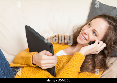 Woman lying on sofa with digital tablet and talking via cell phone Banque D'Images