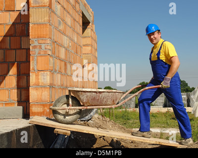 Construction Worker wearing blue dans l'ensemble et le casque, entrer à l'intérieur du bâtiment inachevé mur de brique avec des roues-barrow Banque D'Images