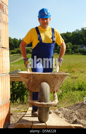 Construction Worker wearing blue coveralls et entrer à l'intérieur du casque de construction de murs en brique non fini avec des roues-barrow Banque D'Images