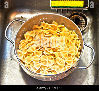 Farfalle fraîchement bouillie, ou Bow Tie, pâtes en drainant passoire dans l'évier de la cuisine avec une éponge jaune vif. Banque D'Images