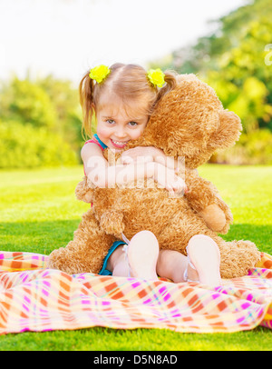 Cute little happy girl hugging Grand ours en peluche brun et s'asseyant sur l'herbe verte prairie, saison printemps Banque D'Images