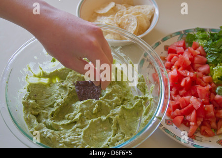 Close up of a hand trempage d'un blue corn chip dans un grand bol de guacamole entouré par la laitue, les tomates et les croustilles de maïs ordinaire Banque D'Images