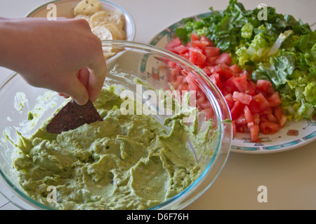 Close up of a young part trempage d'un blue corn chip dans un grand bol de guacamole à côté de croustilles de maïs blanc, tomates et laitue Banque D'Images